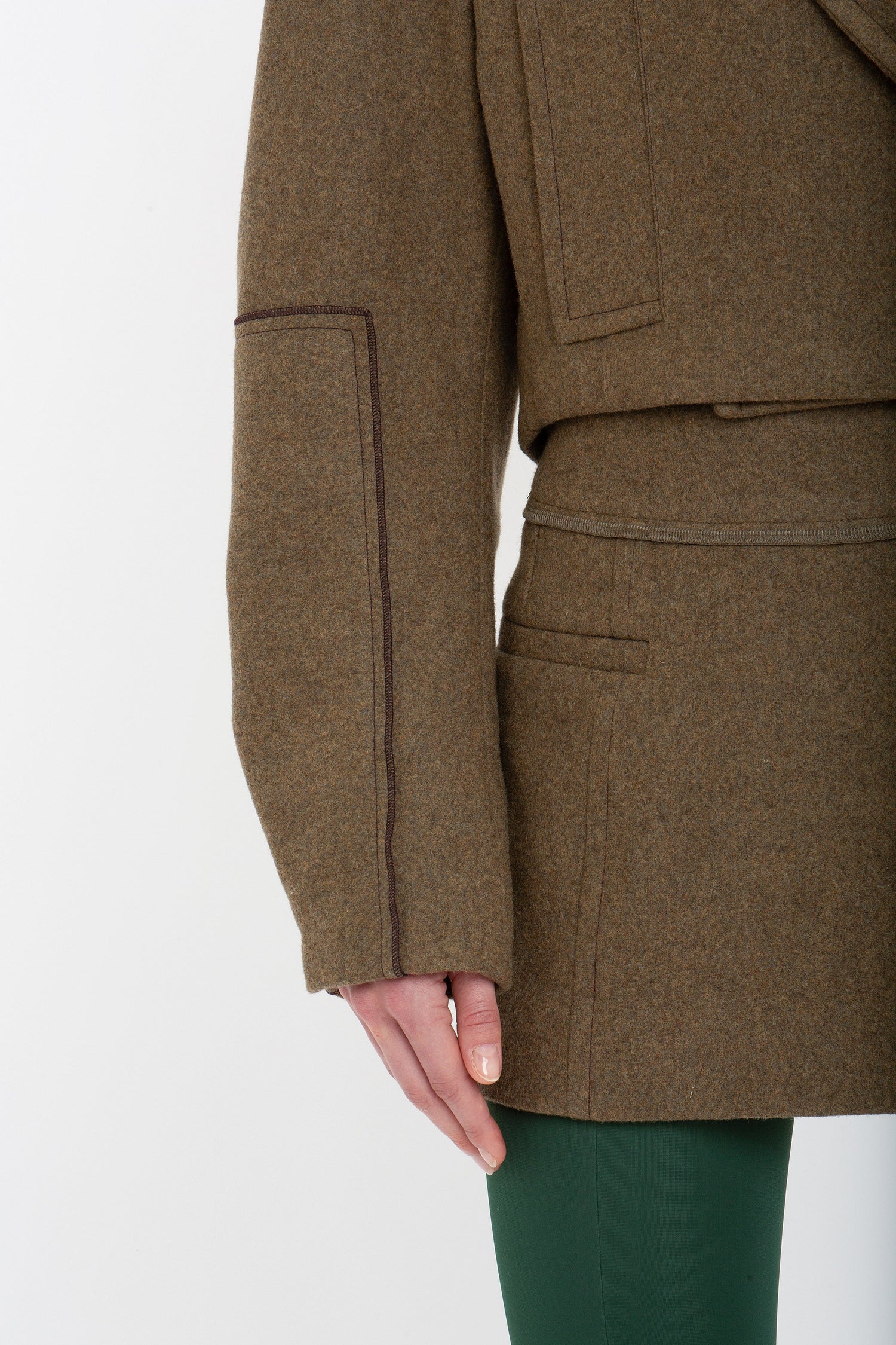 Close-up of a person wearing a brown coat with visible stitching and a Victoria Beckham Tailored Mini Skirt In Khaki, with their left hand resting at their side.