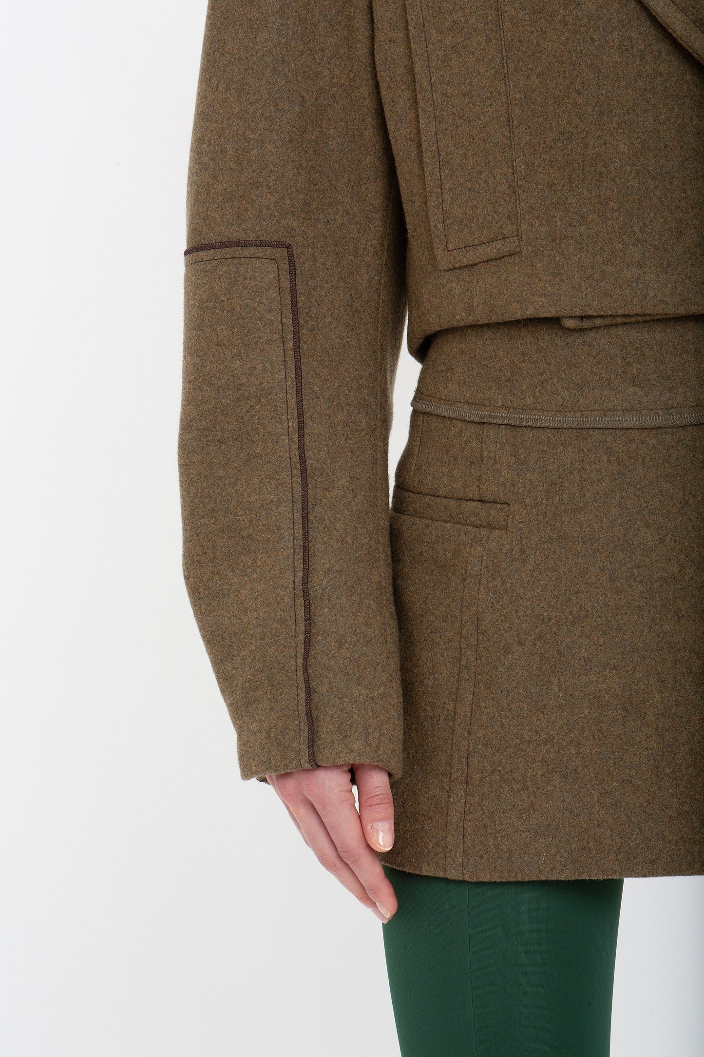 Close-up of a person wearing a Cropped Pea Coat In Khaki by Victoria Beckham with visible seams and green pants, showing the left side of the coat with one arm partially visible.