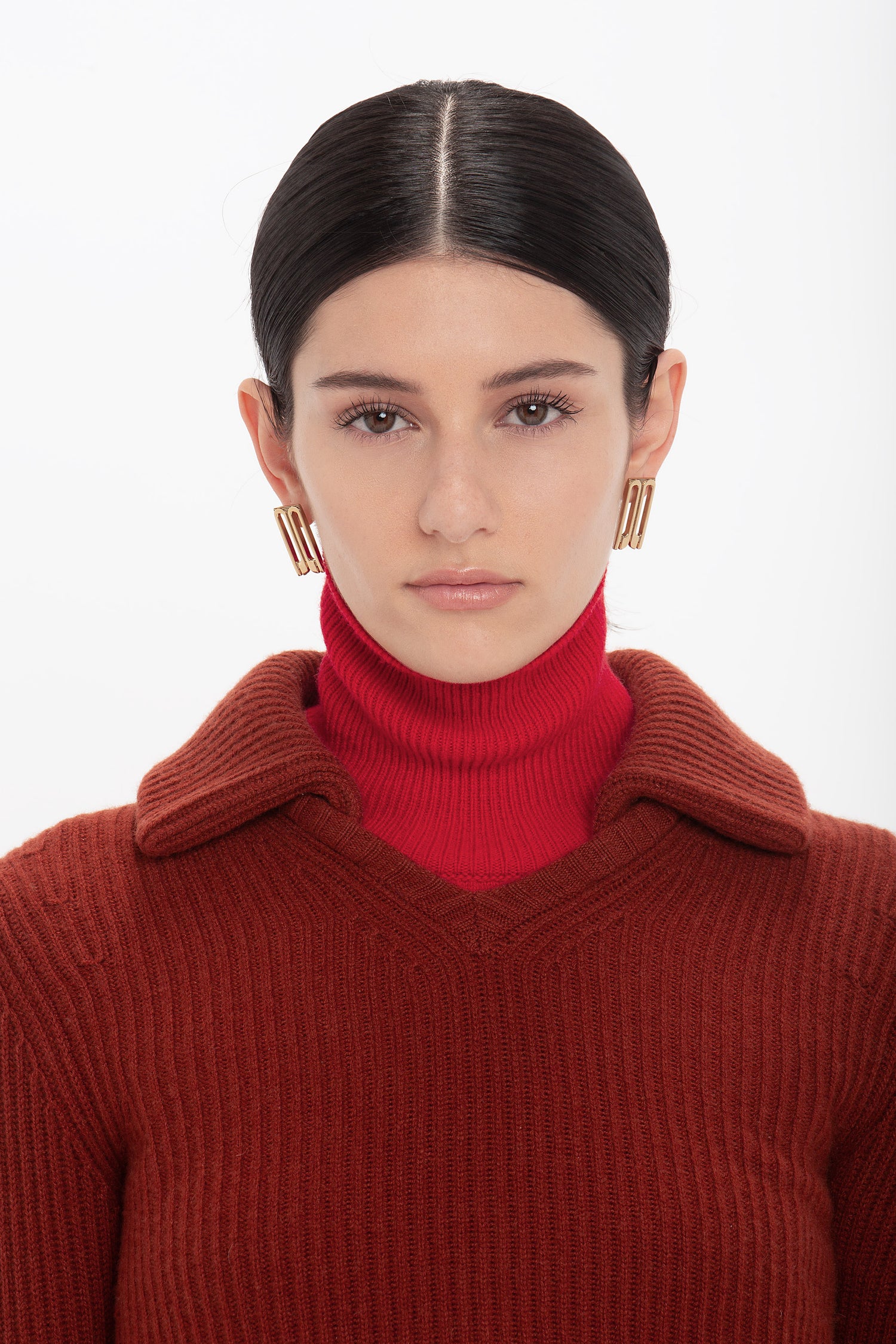 Woman with dark hair slicked back, reminiscent of Victoria Beckham's style, wears a red Double Collared Jumper In Russet by Victoria Beckham and gold hoop earrings, facing the camera.
