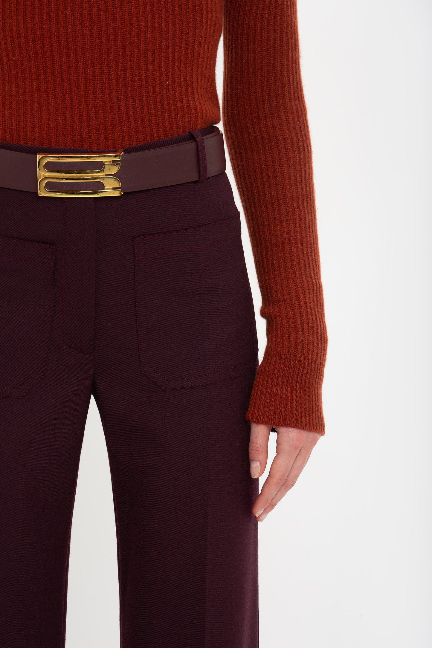 Person wearing a rust-colored, Victoria Beckham Double Collared Jumper In Russet, burgundy pants, and a wide brown belt with a gold buckle, shown from the waist down.