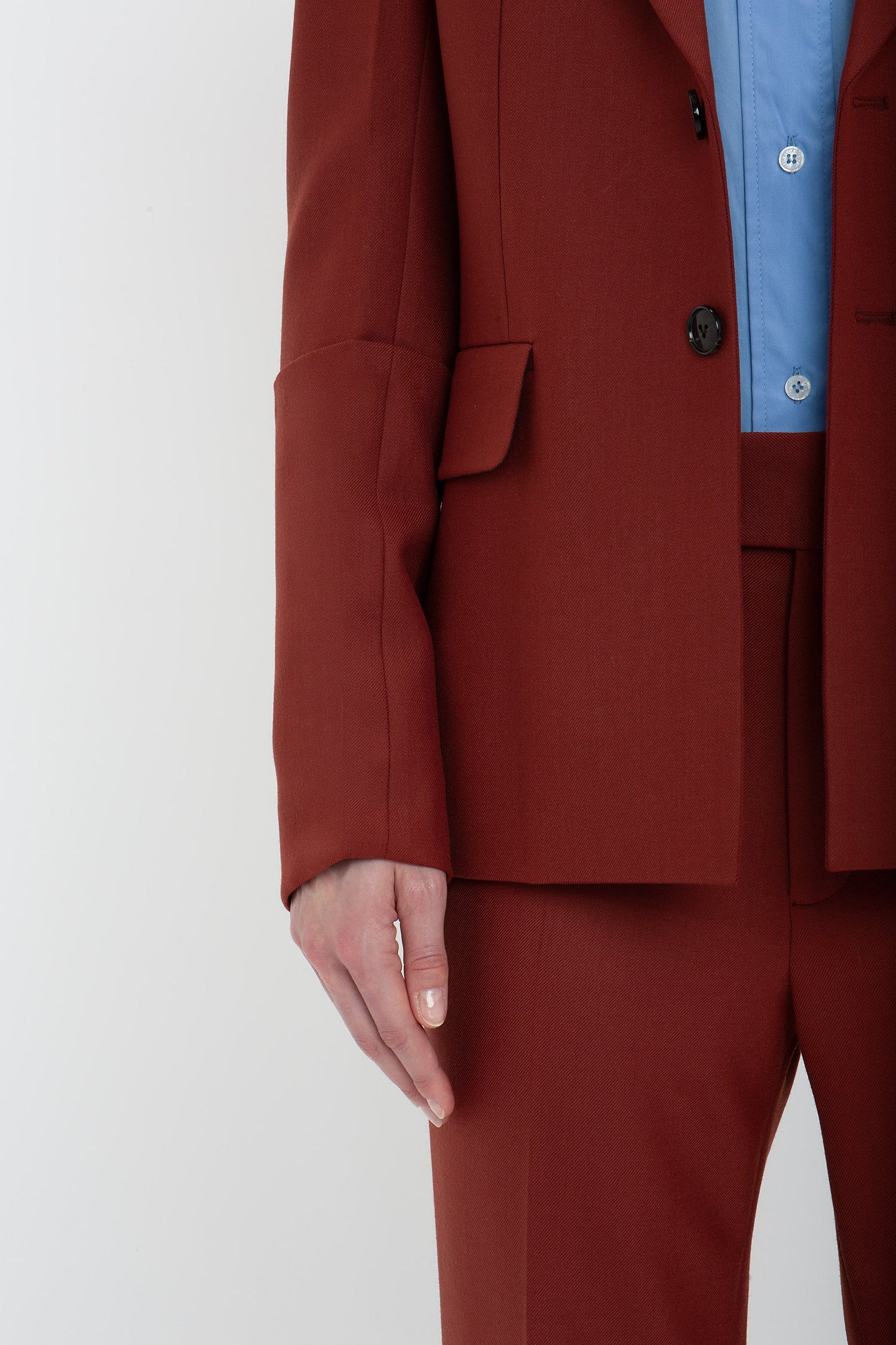 Close-up of a person wearing a Victoria Beckham Sleeve Detail Patch Pocket Jacket In Russet and matching pants in recycled wool, with a light blue shirt underneath. The image shows the person's left arm hanging by their side, accentuating the contemporary detailing of the outfit.
