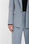 A person wearing a light gray, tailored blazer with a black shirt. The image focuses on the lower part of the Exclusive Sleeve Detail Patch Pocket Jacket In Marina by Victoria Beckham, the sleeve, and the hand.