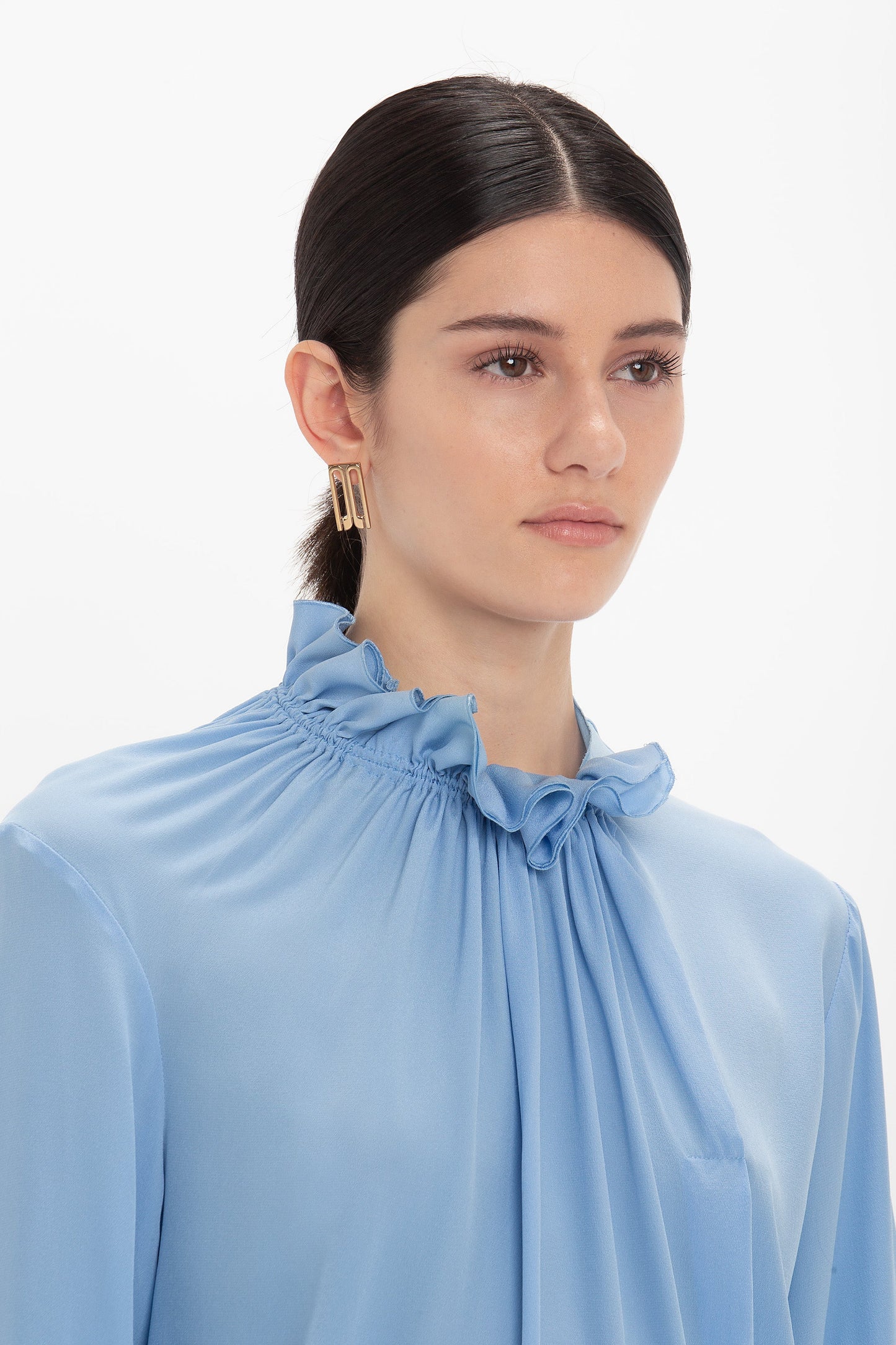 A person with dark hair tied back wearing a light blue crepe de chine fabric, Victoria Beckham Exclusive Ruffle Neck Blouse In Cornflower Blue and gold earrings against a white background.