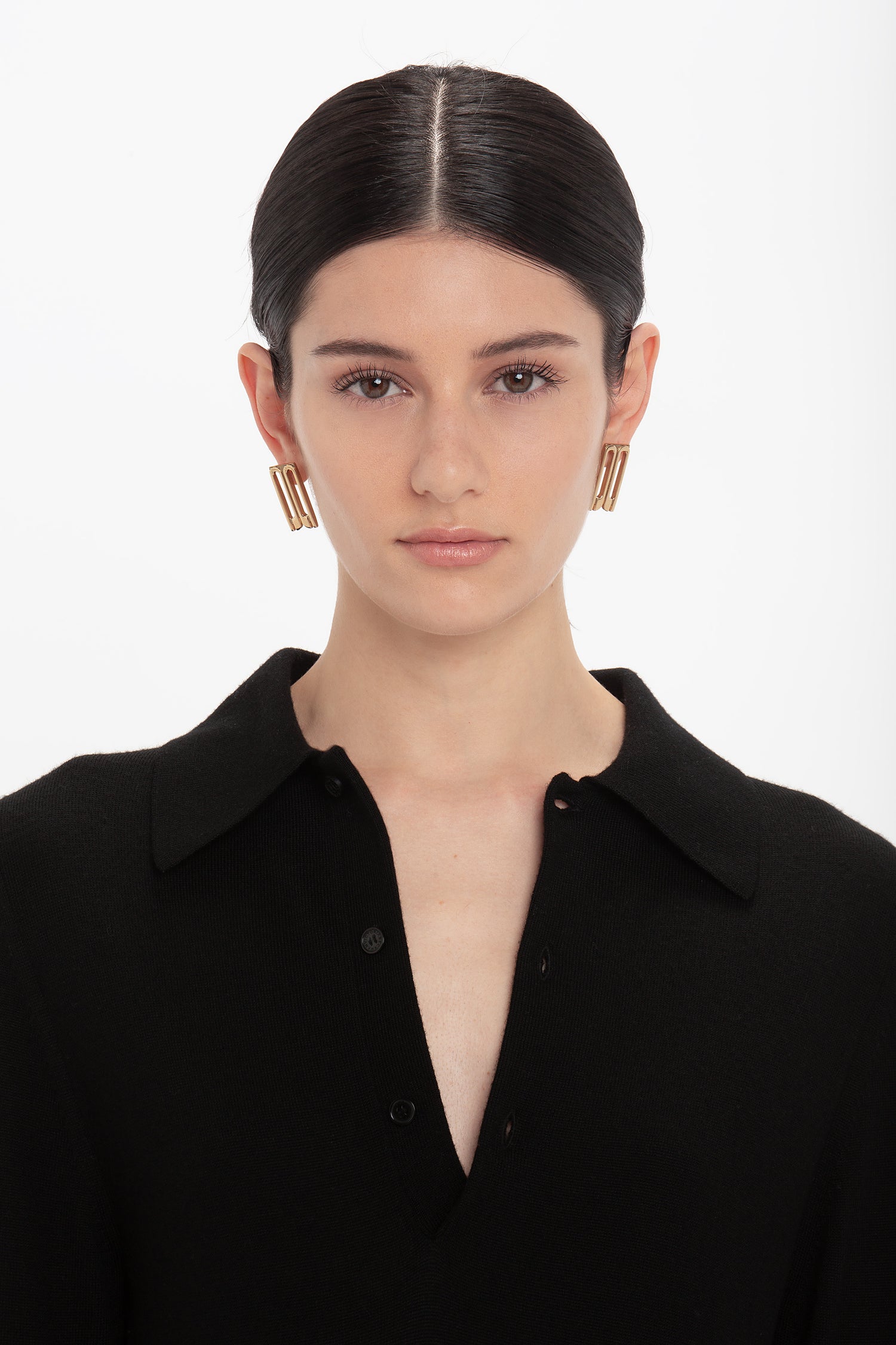 A person with dark hair pulled back wears a black collared shirt and gold rectangular earrings, looking straight ahead with a neutral expression against a plain white background. The asymmetric waist seam of the Henley Shirt Dress In Black by Victoria Beckham adds an intriguing touch to the overall look.