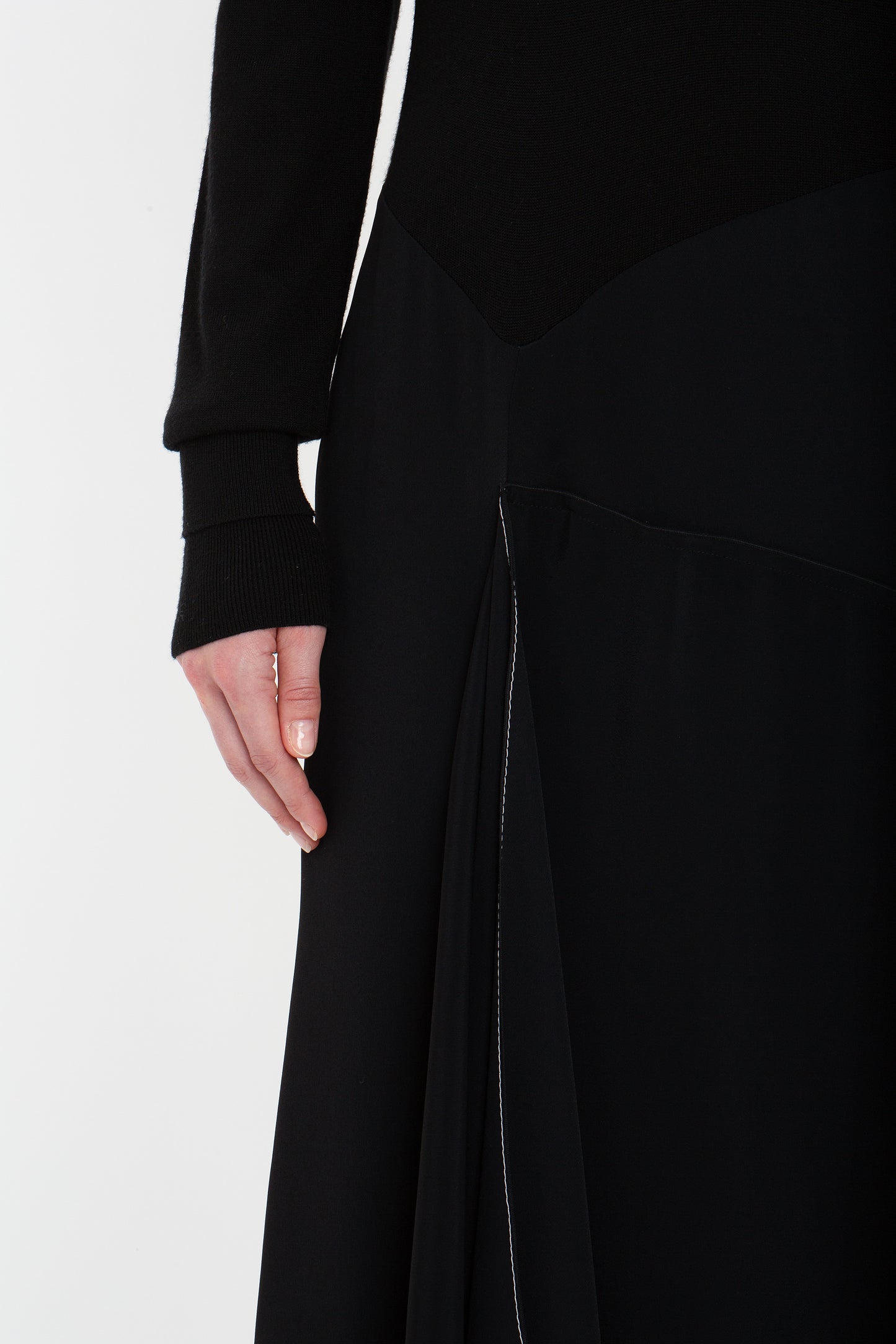 A person in a Henley Shirt Dress In Black by Victoria Beckham, possibly made of merino wool knit, is shown, with one hand visible and resting alongside the dress. The photo focuses on the lower torso and hand area.