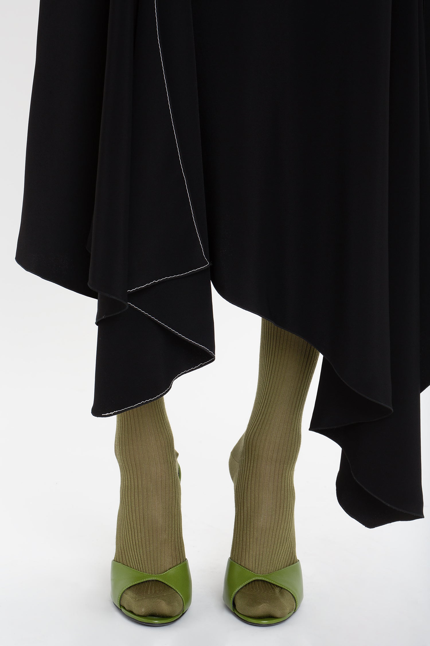 A person wearing a Henley Shirt Dress In Black by Victoria Beckham with an uneven hem and olive-green ribbed socks, paired with olive-green open-toed heels, stands against a white background. The outfit subtly incorporates elements like an asymmetric waist seam that adds a contemporary twist.