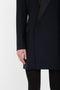 A person stands wearing a dark, long-sleeve Exclusive Fold Shoulder Detail Dress In Midnight by Victoria Beckham and black pants, reminiscent of sophisticated evening wear, with one hand visible by their side against a plain white background.