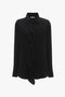 The Victoria Beckham Asymmetric Ruffle Blouse In Black is a classic black long-sleeve blouse with an asymmetric ruffle front detail and a collared neckline, crafted from fluid silk.