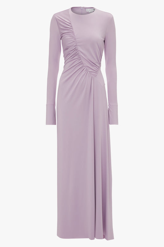 A long-sleeved, lavender-colored dress crafted from stretch jersey with a cinched side detail and a floor-length hemline exudes understated glamour against a plain background. The Victoria Beckham Ruched Detail Floor-Length Gown In Petunia transforms elegance into an effortless statement.