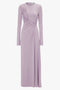 A long-sleeved, lavender-colored dress crafted from stretch jersey with a cinched side detail and a floor-length hemline exudes understated glamour against a plain background. The Victoria Beckham Ruched Detail Floor-Length Gown In Petunia transforms elegance into an effortless statement.