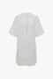 A loose, white Frame Cut-Out T-Shirt Dress In White crafted from bonded linen jersey with short sleeves and a navel-grazing keyhole neckline. The bottom features a layered hem detail for added elegance. This sophisticated piece by Victoria Beckham combines casual comfort with chic style.