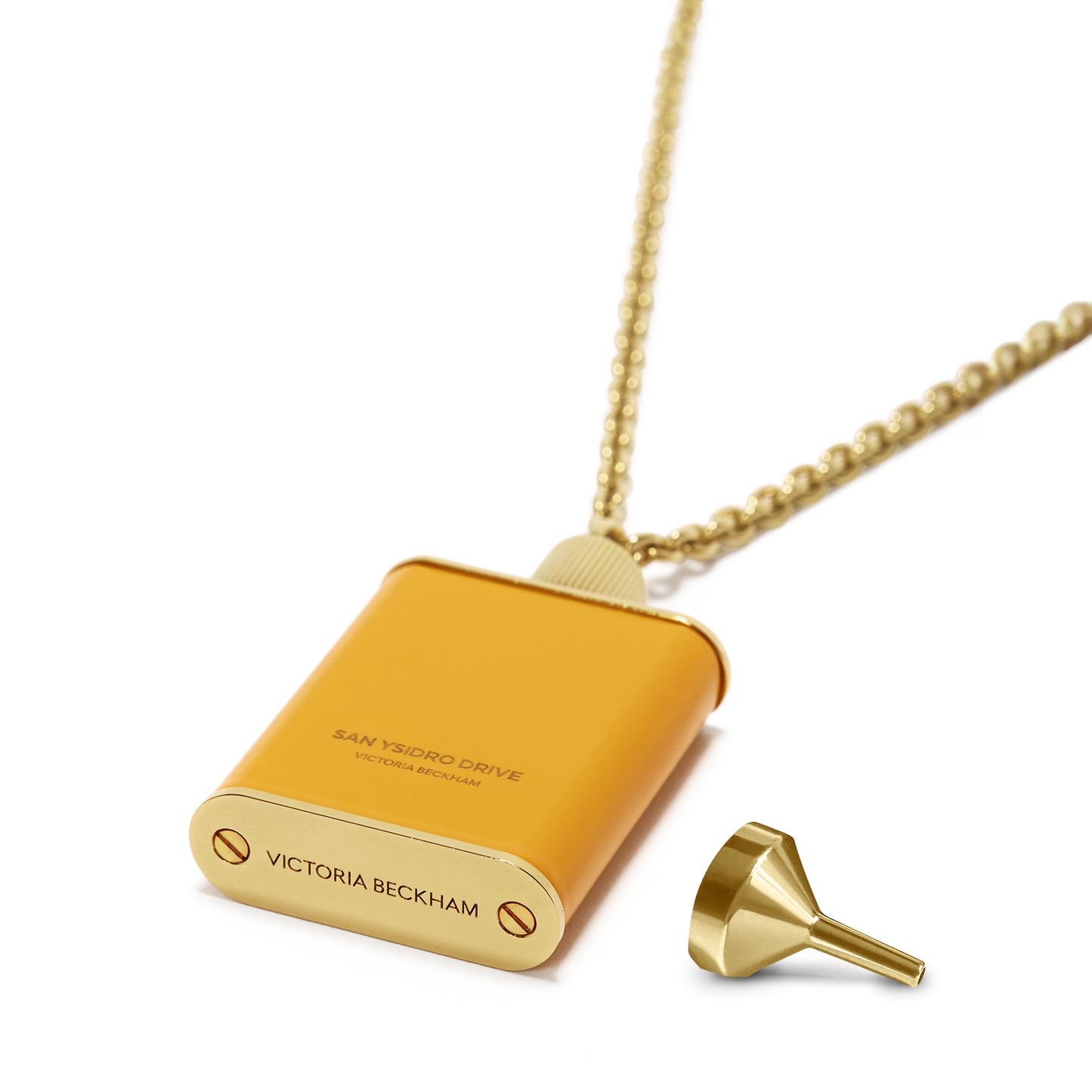 A gold brushed brass pendant on a chain features the label "San Ysidro Drive Victoria Beckham" and "Victoria Beckham" engraved. The Perfume Bottle Necklace In San Ysidro Drive by Victoria Beckham is placed beside it.