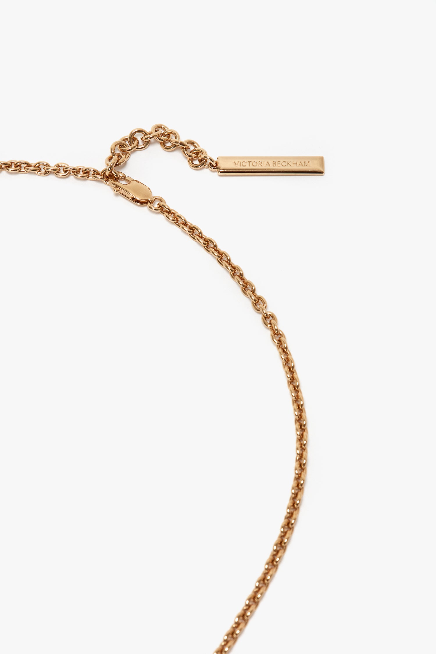 Close-up of a gold brushed brass chain necklace with a small rectangular tag that reads "Victoria Beckham." The chain is secured with a round clasp, showcasing distinct Victoria Beckham branding.