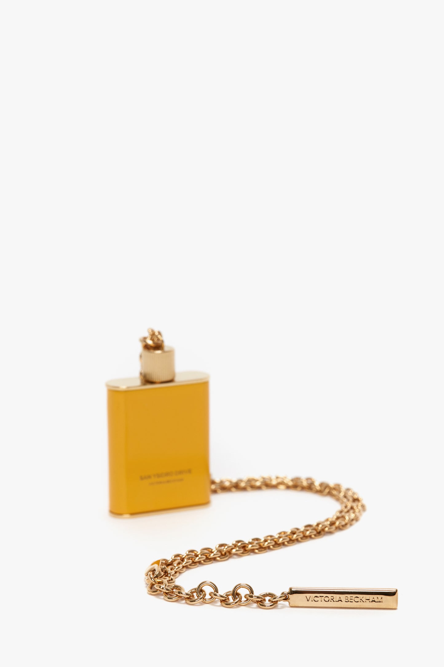A golden, rectangular lighter with an attached chain and a tag inscribed with "Victoria Beckham" exudes elegance. The gold brushed brass finish perfectly complements the Victoria Beckham branding, making it a luxurious accessory.