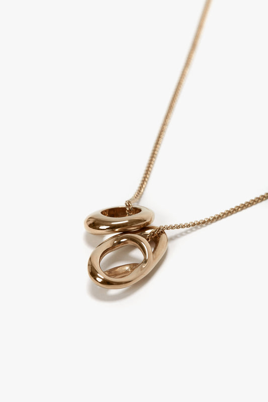 A light gold necklace featuring two interlocking oval-shaped pendants on a fine chain with a T-bar closure, displayed against a white background. The Victoria Beckham Exclusive Abstract Charm Necklace in Light Gold embodies timeless elegance suitable for any occasion.