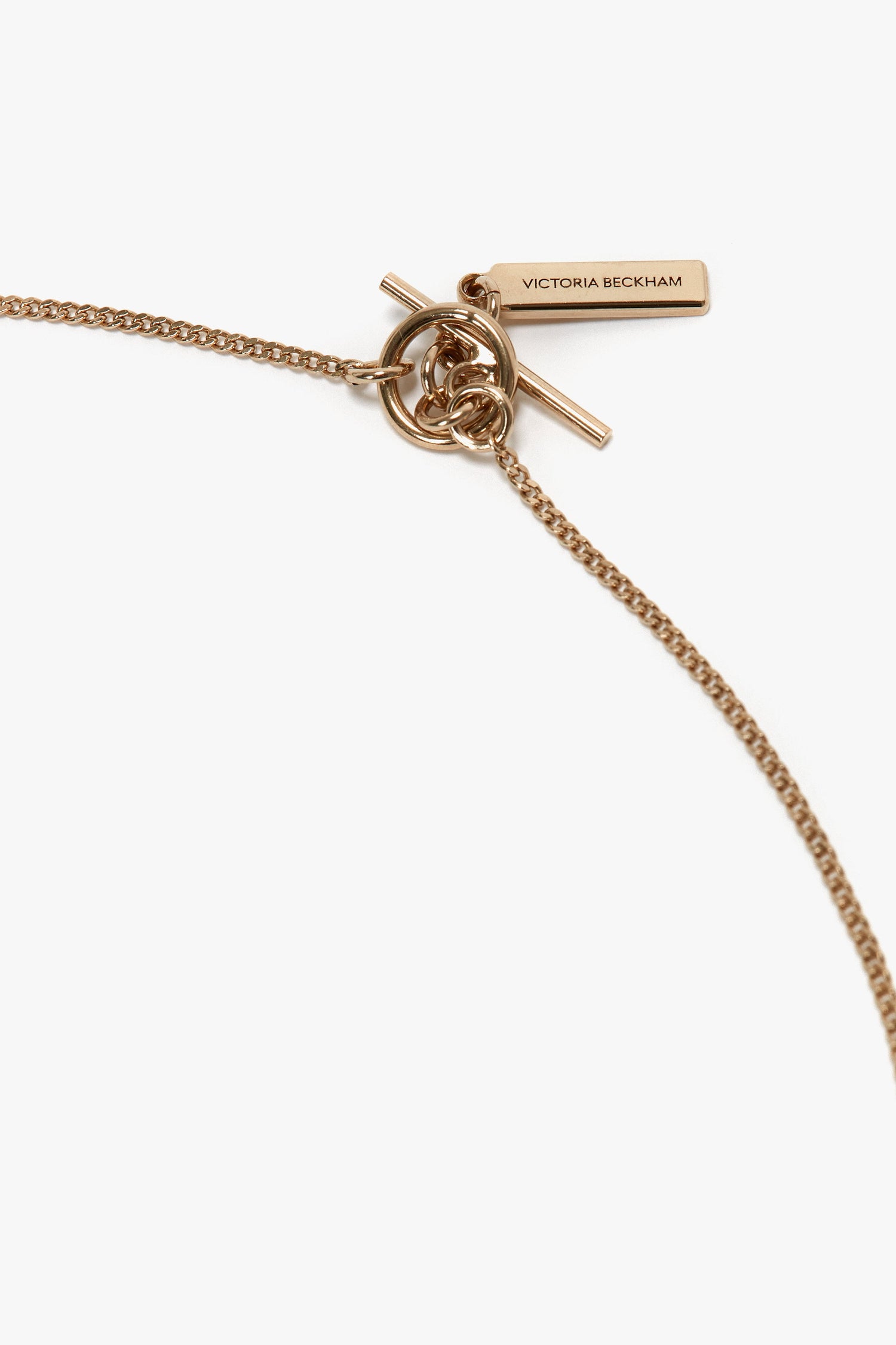 A close-up of an Exclusive Abstract Charm Necklace In Light Gold features a T-bar closure and a rectangular tag engraved with "Victoria Beckham," exuding the elegance of an abstract charm necklace.