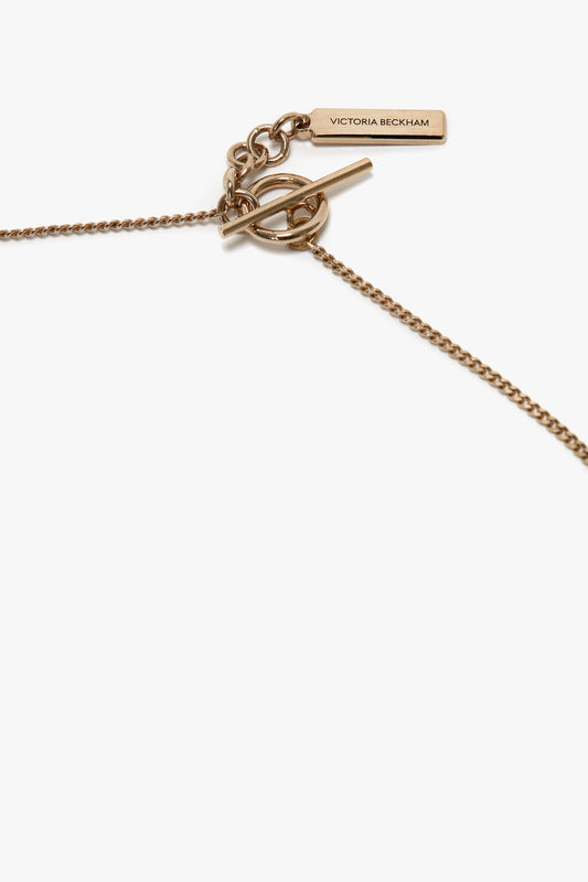 A close-up of an Exclusive Body Charm Necklace In Light Gold, featuring a delicate chain design and a toggle clasp engraved with "Victoria Beckham" on a rectangular tag. Crafted from 100% brass, this elegant piece adds a touch of sophistication to any outfit.