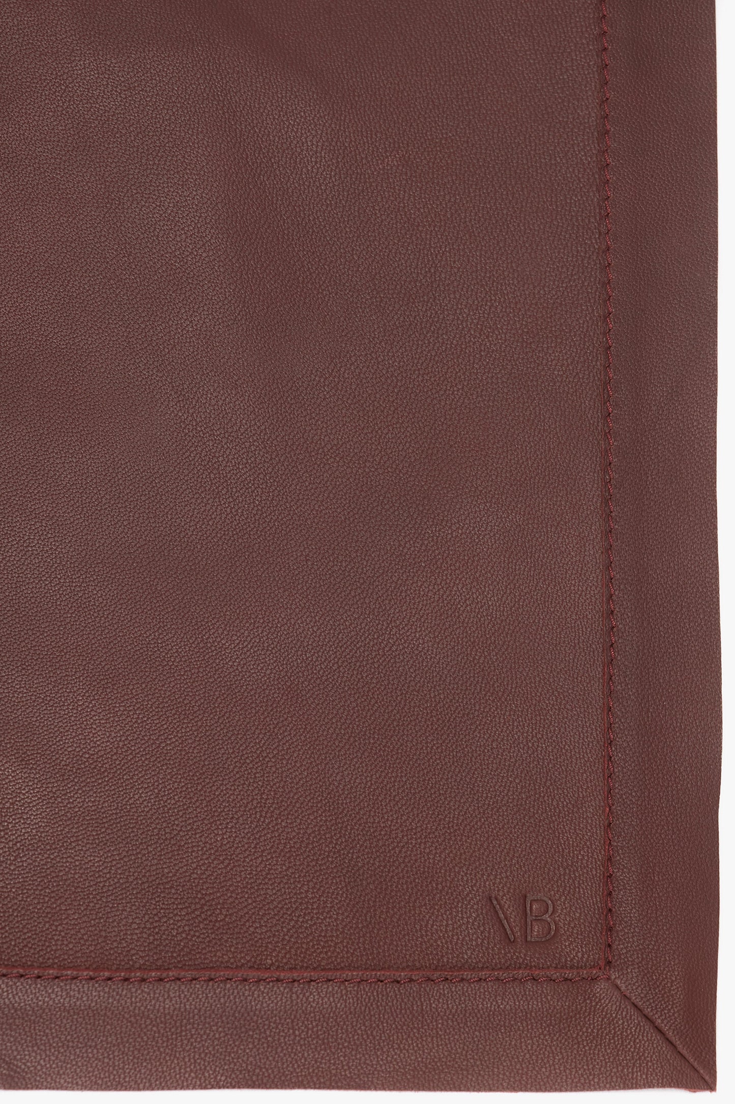 Close-up view of a corner of a maroon leather item with visible stitching and a small debossed logo in the bottom right, epitomizing the blend of masculine tailoring and refined craftsmanship, this Victoria Beckham Leather Scarf In Bordeaux showcases exquisite detail.