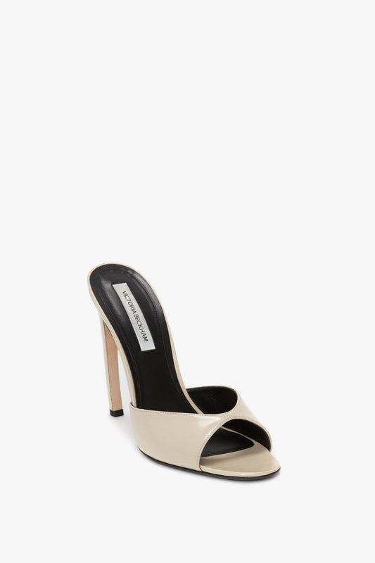 A beige high-heeled open-toe mule with a black interior and a thin heel, featuring luxurious calf leather and a chic V cut design, the Classic Mule In Macadamia calf Leather by Victoria Beckham.