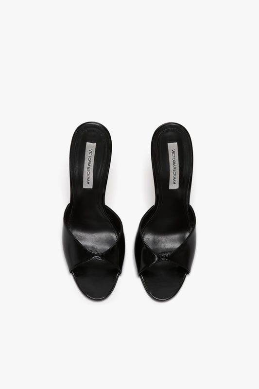 A pair of black, open-toe slide sandals with crisscross straps and a seductive curved heel, viewed from above. Labels inside the shoes read "Victoria Beckham Classic Mule In Black Calf Leather.