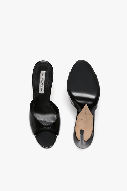A pair of Classic Mule In Black Calf Leather by Victoria Beckham with one shoe showing the top view and the other showing the sole, crafted in luxury calf leather. The insole has a visible white label, exuding both elegance and comfort.