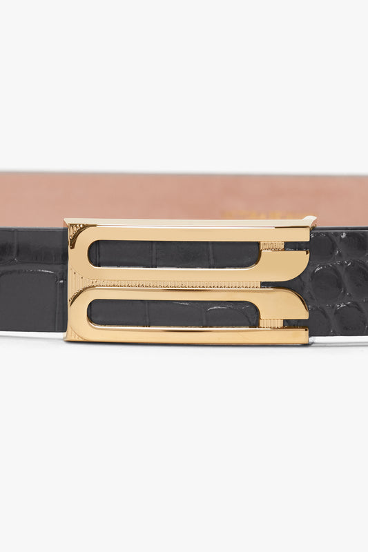 A close-up image of a **Frame Belt In Slate Grey Croc Embossed Calf Leather** with a gold buckle featuring a decorative, geometric design by **Victoria Beckham**.