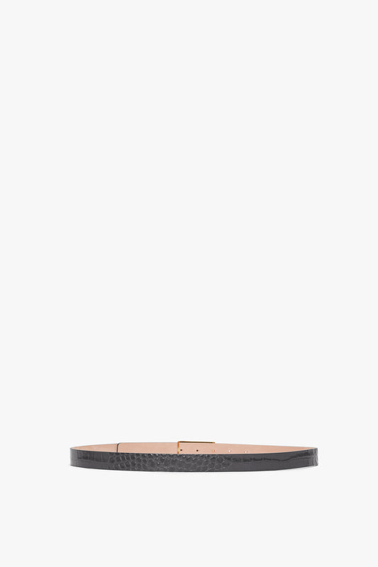 A thin black Victoria Beckham Frame Belt In Slate Grey Croc Embossed Calf Leather features a subtle pattern and a rectangular, metallic gold buckle laid flat against a white background.