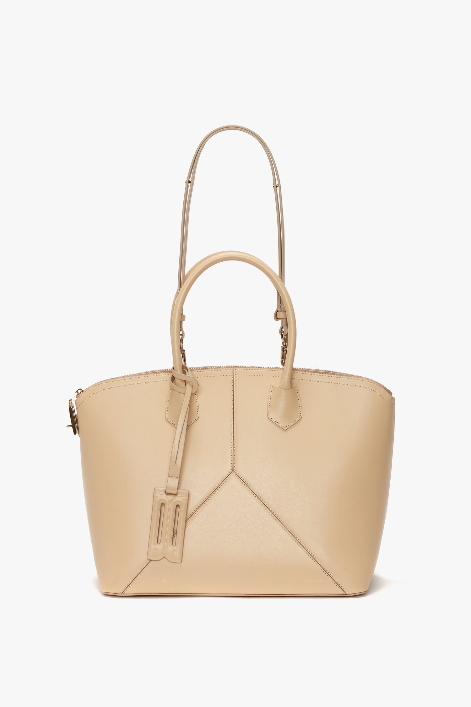 The Victoria Beckham Victoria Bag In Sesame Leather is a tan leather handbag with dual handles and an adjustable, detachable shoulder strap, featuring angular stitched leather panels on the front.