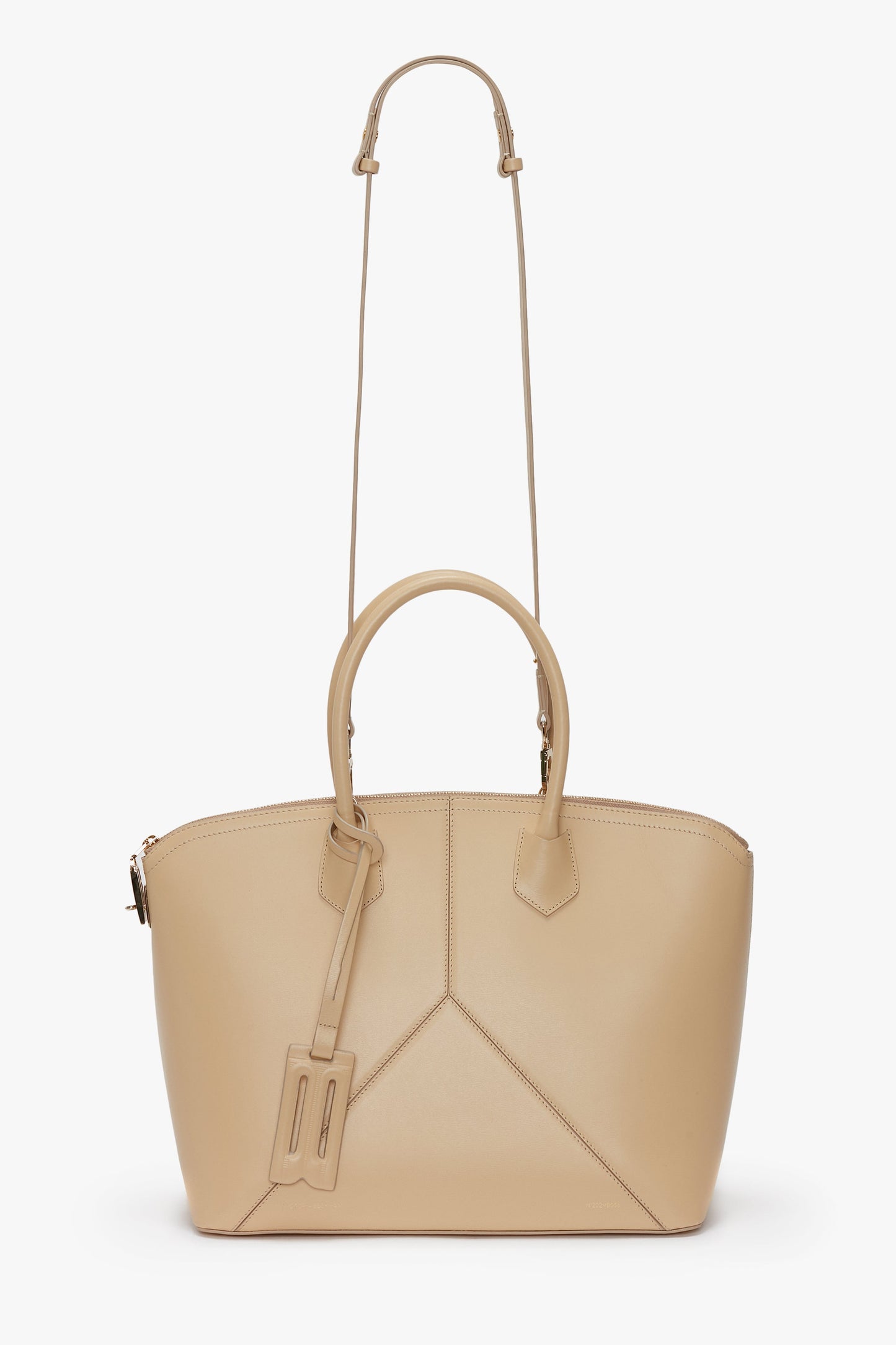 The Victoria Beckham Victoria Bag In Sesame Leather features dual top handles, leather panels, an adjustable shoulder strap, and a luggage tag attached.
