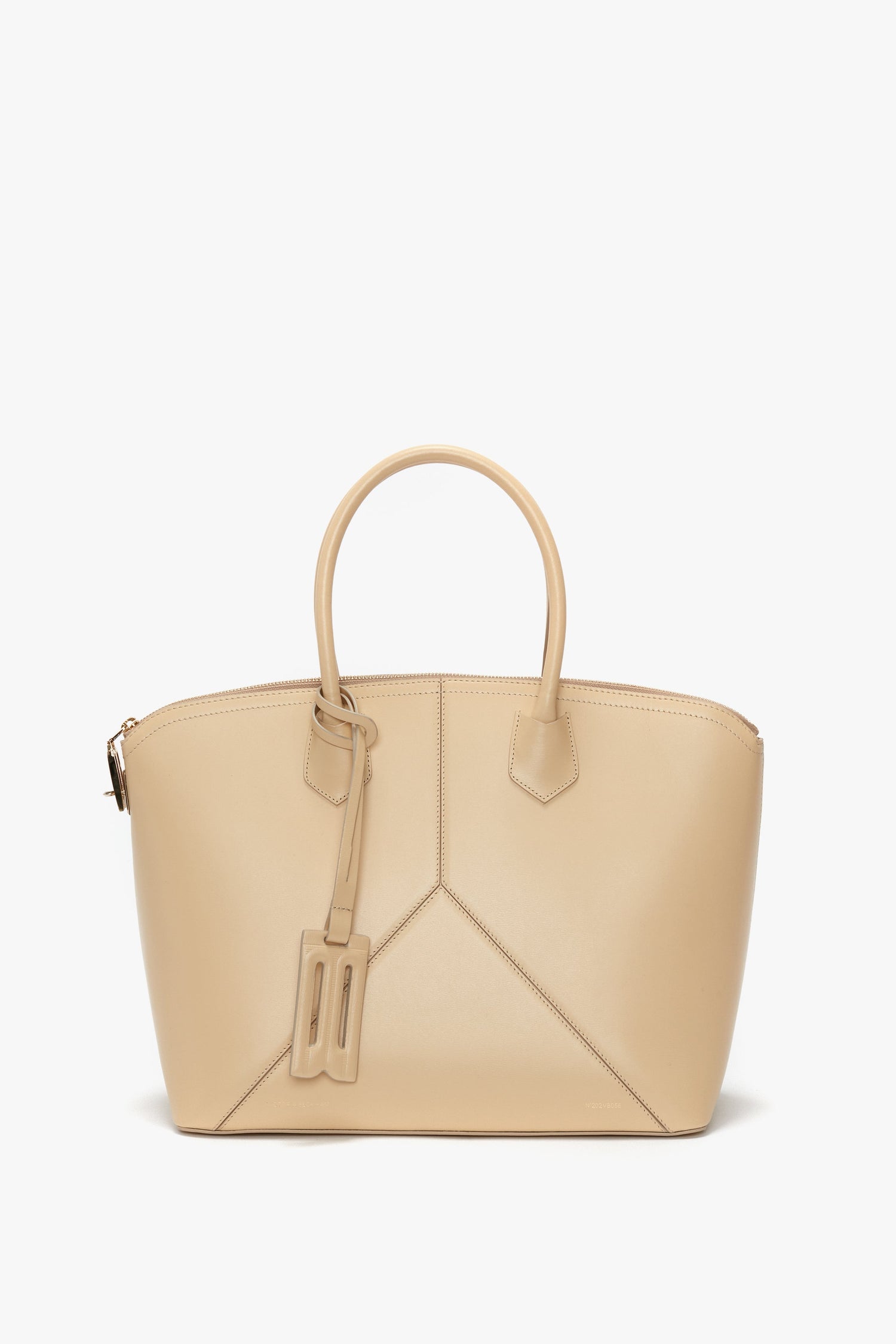 A beige Victoria Beckham Victoria Bag In Sesame Leather with leather panels, curved handles, and a decorative tag hanging from one handle features an adjustable strap for versatility.