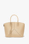 A beige Victoria Beckham Victoria Bag In Sesame Leather with leather panels, curved handles, and a decorative tag hanging from one handle features an adjustable strap for versatility.