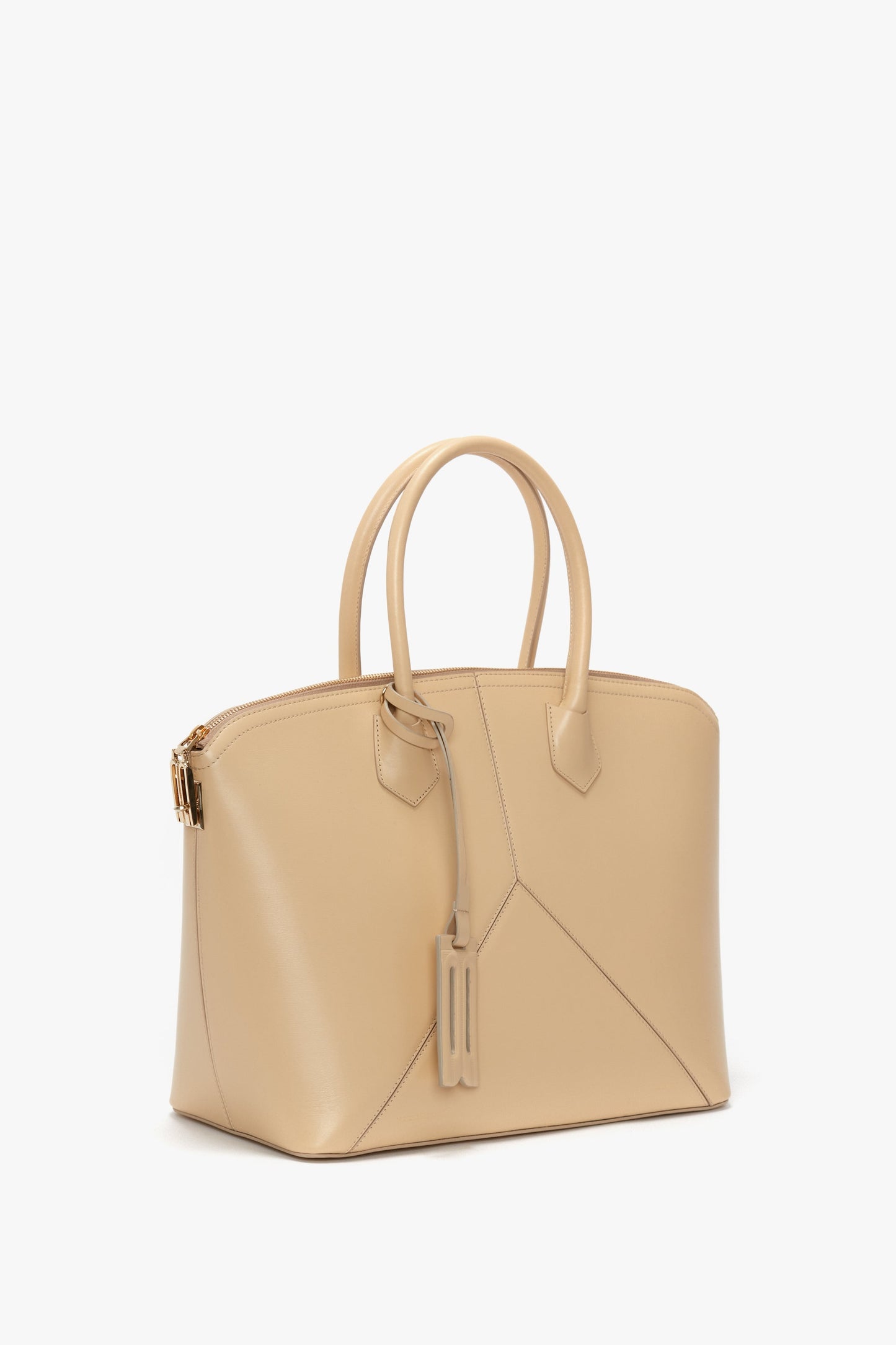 A beige Victoria Beckham Victoria Bag In Sesame Leather with a geometrical design, top handles, leather panels, a zipper closure, and a luggage tag attached.