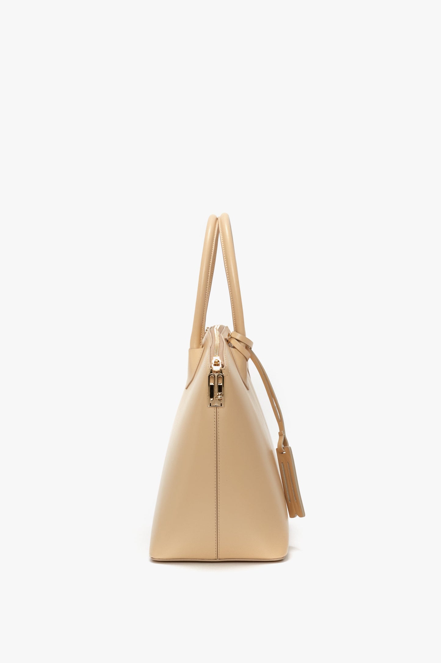 Side view of the Victoria Beckham Victoria Bag In Sesame Leather, a beige handbag with double handles, leather panels, and a tassel decoration, featuring a gold zipper closure against a white background.