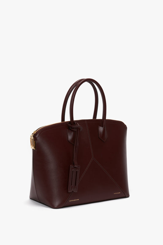 The Victoria Beckham Victoria Bag In Burgundy Leather is a dark brown leather handbag with two handles, a luggage tag, and gold hardware. Featuring a zippered top, structured shape, and padlock closure for added security, this elegant piece ensures both style and function in every detail.