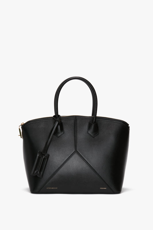 The Victoria Beckham Victoria Bag In Black Leather is a chic leather handbag with dual handles and a zippered top closure. It features a geometric stitching design, a luggage tag attached to one handle, and an elegant debossed charm for added sophistication.