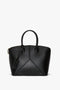 The Victoria Beckham Victoria Bag In Black Leather is a chic leather handbag with dual handles and a zippered top closure. It features a geometric stitching design, a luggage tag attached to one handle, and an elegant debossed charm for added sophistication.