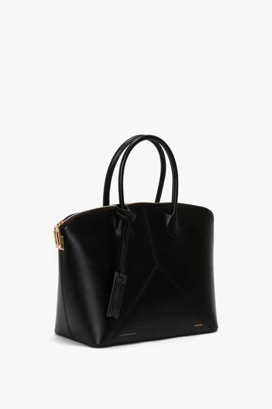 The Victoria Beckham Victoria Bag In Black Leather is a black leather hold-all handbag with a gold zipper, two handles, and a small attached tag against a white background.