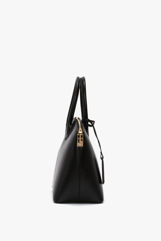 Side view of the Victoria Beckham Victoria Bag In Black Leather, a black hold-all with gold-tone zipper and hardware, featuring a structured design, two handles, and a leather debossed charm.