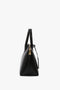 Side view of the Victoria Beckham Victoria Bag In Black Leather, a black hold-all with gold-tone zipper and hardware, featuring a structured design, two handles, and a leather debossed charm.