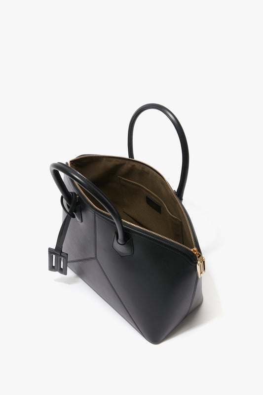The Victoria Beckham Victoria Bag In Black Leather is a black leather hold-all featuring a gold zipper that's partially open, revealing a brown interior lining. The bag has black handles, a strap with a buckle, and is adorned with a stylish leather debossed charm.