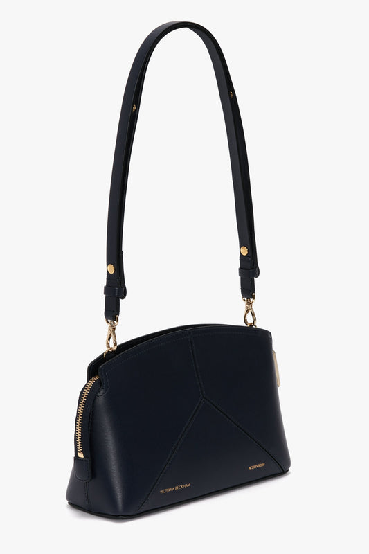 A black leather handbag with gold accents, a thick strap, and geometric stitching on the front. The luxuriously textured Exclusive Victoria Crossbody Bag In Navy Leather by Victoria Beckham features a zip closure and finely crafted leather panels.