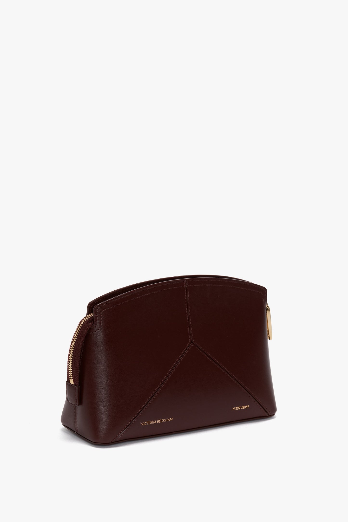 A dark brown, semi-curve shaped Victoria Crossbody Bag In Burgundy Leather, crafted from rich burgundy leather by Victoria Beckham, featuring gold-toned zippers and minimalistic branding embossed on the front.
