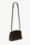 The Victoria Beckham Exclusive Victoria Crossbody Bag In Brown Leather is a dark brown calf leather bag with gold-tone hardware, an adjustable strap, and a zip closure.