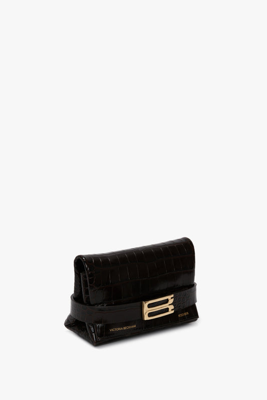 A small, black, crocodile-textured Mini B Pouch Bag In Croc Effect Espresso Leather crafted from calf leather with a gold buckle and the name "Victoria Beckham" inscribed on it. It features a stylish, detachable crossbody strap for versatility.