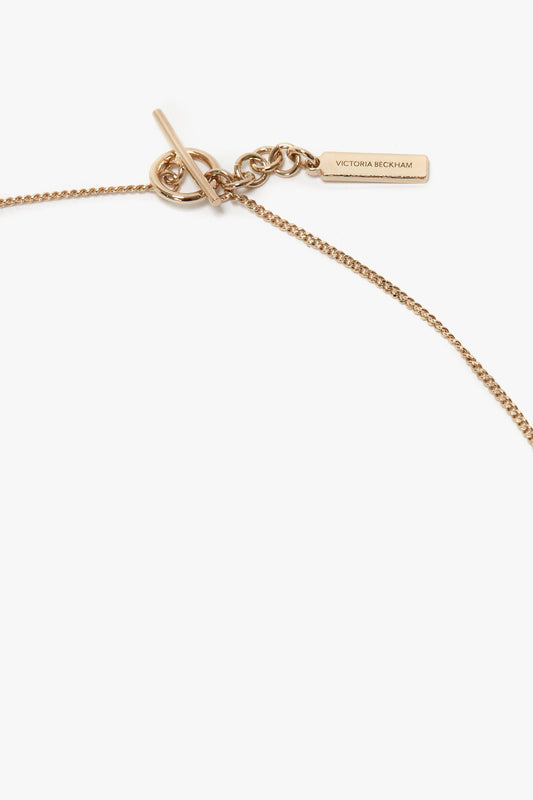 A delicate light gold chain necklace with a bar and circle clasp. It features a small rectangular tag engraved with "Victoria Beckham." The background is plain white. Made from 100% brass, this Exclusive Resin Pendant Necklace In Light Gold-White by Victoria Beckham adds a touch of elegance to any outfit.