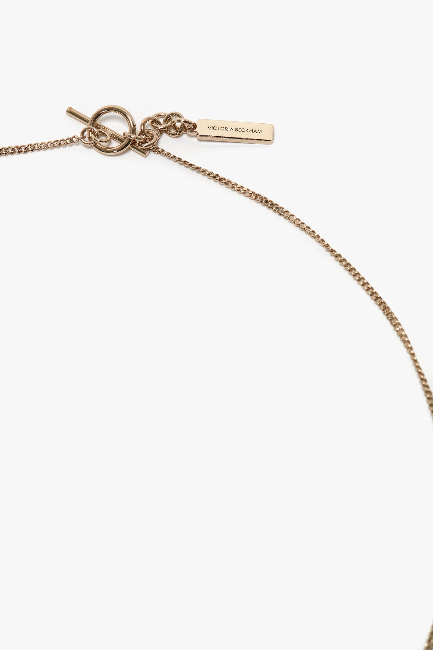 A close-up of a delicate gold chain necklace with a toggle clasp, featuring a small tag inscribed with "Victoria Beckham." This exquisite piece, the Exclusive Resin Pendant Necklace In Light Gold-Orange by Victoria Beckham, made in Italy, exudes elegance and sophistication.