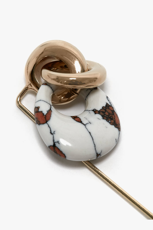 A Victoria Beckham Exclusive Resin Charm Brooch In Light Gold-White featuring a marble-patterned circular stone intertwined with a metallic loop, attached to a long, straight pin. This exquisite piece boasts a light gold/white design and is crafted with the finest details, making it an elegant addition to your collection.