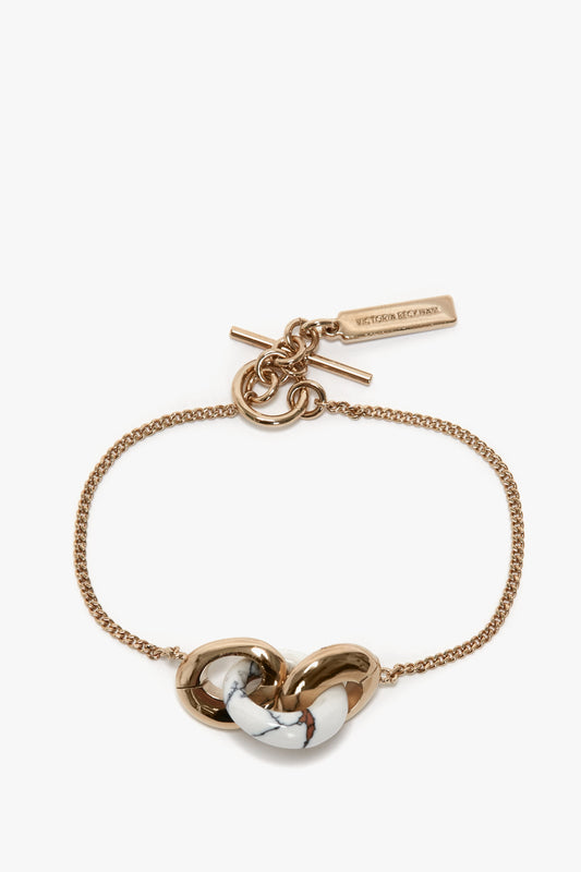 An Exclusive Resin Charm Bracelet In Light Gold-White made in Italy, featuring a gold chain with a light gold/white stone and gold knot centerpiece, complemented by a bar and ring toggle clasp. It boasts a rectangular tag engraved with "Victoria Beckham", embodying elegance.