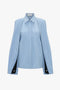 A Victoria Beckham Pleat Detail Raglan Shirt In Oxford Blue with a unique cape-like design and contemporary silhouette. The sleeves have black inset detailing near the cuffs, adding a touch of elegance. Featuring a classic collar and concealed button-front closure, this shirt exudes a relaxed attitude.