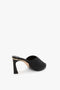 A V Cut Mule In Black Raffia by Victoria Beckham featuring an asymmetrical V cut and sleek black heel, complemented by an open back design on a white background.